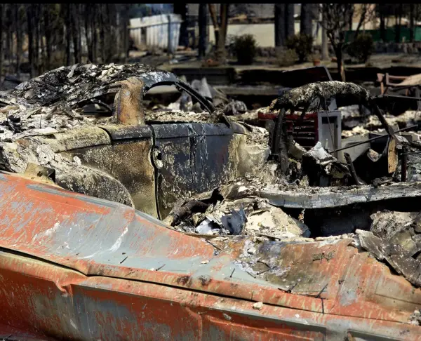 The bed of a pickup in the foreground, the shell of a car and a collapsed house behind it.